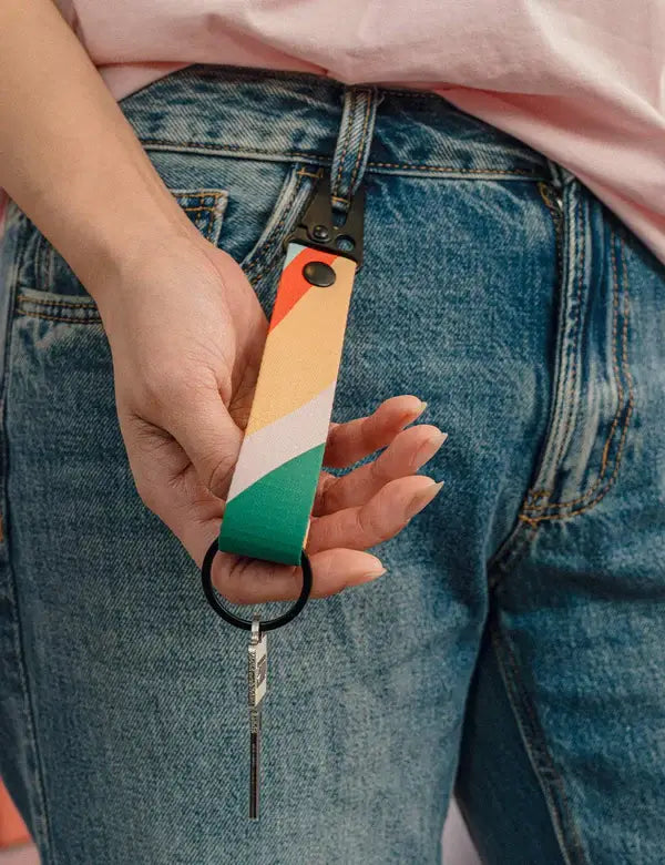 Eine Hand hält den Notabag-Schlüsselanhänger „Flow“ – einen farbenfrohen Schlüsselanhänger in Orange, Pfirsich, Weiß und Grün von Notabag –, der an der Gürtelschlaufe einer blauen Jeans befestigt ist und an dem Schlüssel an einem schwarzen Ring hängen.