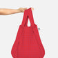 Eine Hand hält die Notabag | Wandelbare Tasche und Rucksack „Beeren“ von Notabag, die sich durch ein wiederverwendbares rotes Design mit kleinem Karomuster und einem weißen Seitenetikett auszeichnet und sich vor einem schlichten, hellen Hintergrund mühelos von einer Einkaufstasche in einen Rucksack verwandeln lässt.