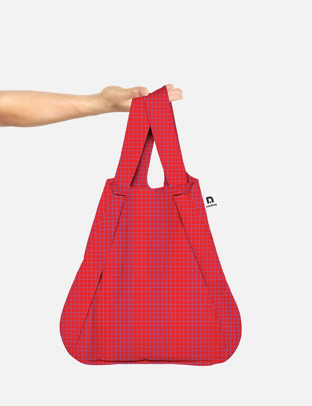 Eine Hand hält die Notabag | Wandelbare Tasche und Rucksack „Beeren“ von Notabag, die sich durch ein wiederverwendbares rotes Design mit kleinem Karomuster und einem weißen Seitenetikett auszeichnet und sich vor einem schlichten, hellen Hintergrund mühelos von einer Einkaufstasche in einen Rucksack verwandeln lässt.