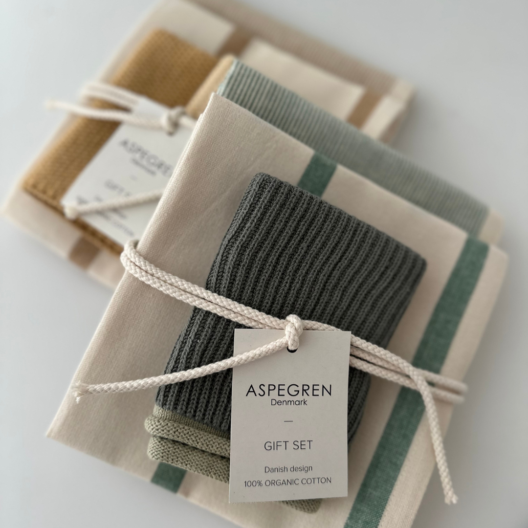 Zwei ordentlich gefaltete Geschenksets von Aspegren Denmark | „Grünes Küchentuch 3er-Geschenkset“, die jeweils gemusterte Küchentücher und einen Waschlappen enthalten, mit einer weißen Schnur zusammengebunden und mit der Aufschrift „100 % Bio-Baumwolle, dänisches Design“ versehen sind.
