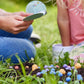 Zwei Personen sitzen auf sattgrünem Rasen und pflanzen begeistert die bunten Blumenmurmeln aus der Saatgut | Blumenmurmeln „Naturabenteuer“ 100er-Dose von Stadtgärtner in die Erde – ein echtes Naturabenteuer für Kinder! Während eine Person die Anleitung in der Hand hält, verfolgt die andere gespannt das bunte Treiben. Im Hintergrund sind praktische Saatgutbehälter zu sehen – ein nachhaltiges Geschenk von Stadtgärtner, das Kindern auf spielerische Weise Freude an der Natur vermittelt.