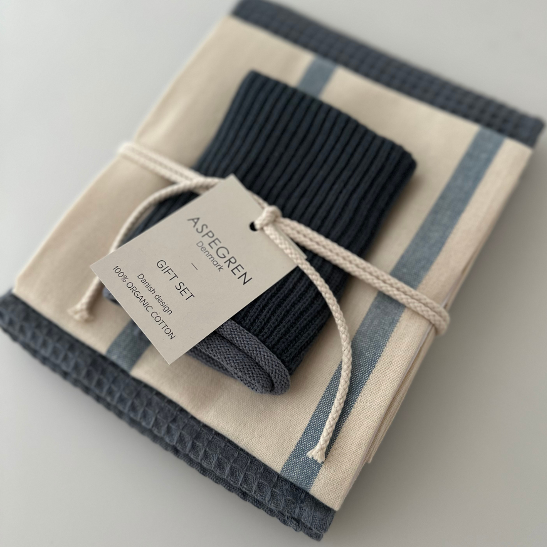 Ein sorgfältig gefaltetes 3-teiliges Set hochwertiger Küchentücher von Aspegren Denmark in tiefen nordischen Blautönen, mit einer weißen Schnur zusammengebunden und mit der Aufschrift „Aspegren Denmark Geschenkset, 100 % Bio-Baumwolle“ versehen. Diese stilvollen Küchentextilien verleihen jeder Küche einen Hauch von Eleganz und sind zugleich praktisch und umweltfreundlich.