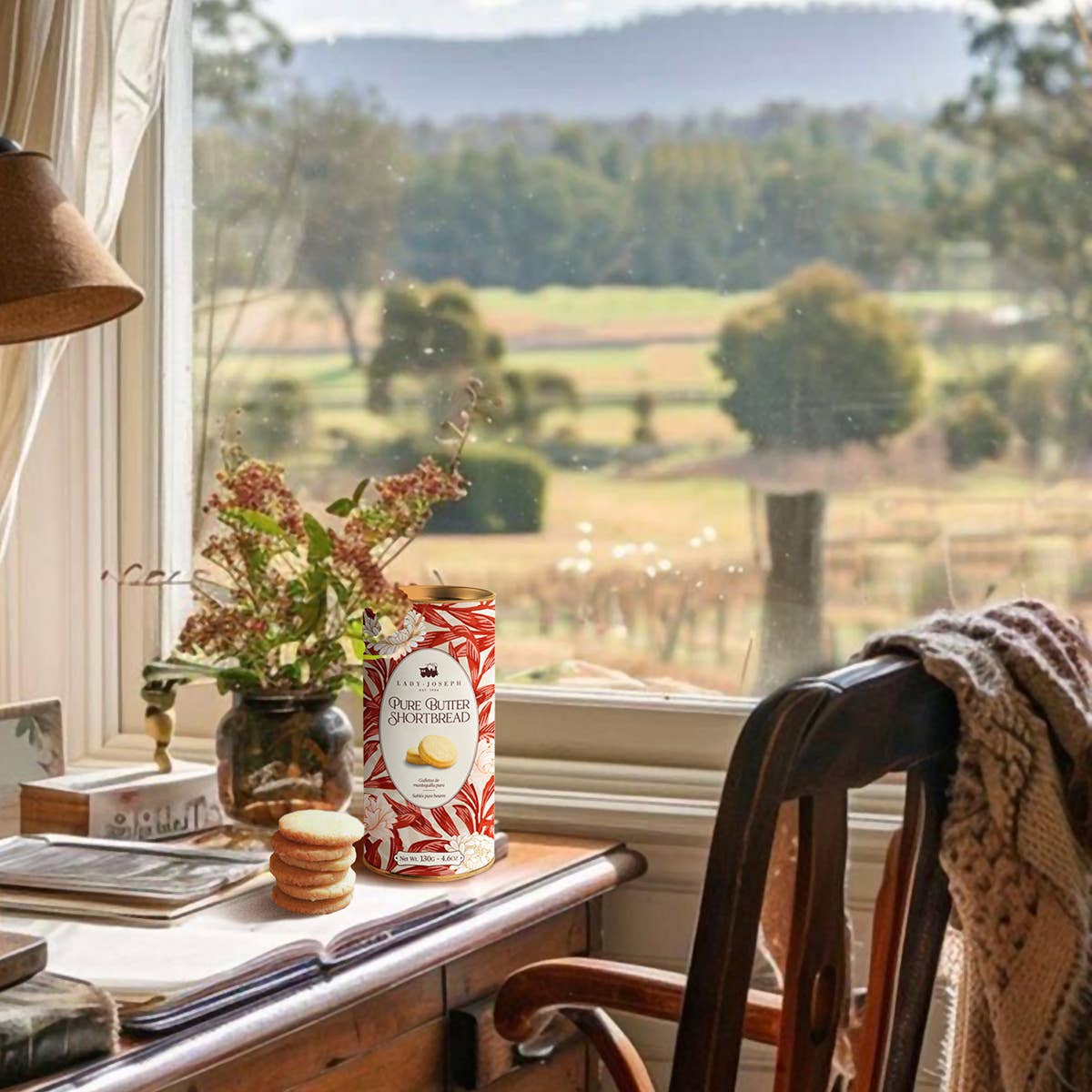 Ein gemütlicher Schreibtisch an einem Fenster mit Blick auf die Landschaft. Darauf liegen Lady Joseph Doppel-Butter-Shortbread 130g, eine Vase mit Trockenblumen, eine Lampe, ein Notizbuch und eine über den Stuhl drapierte Strickdecke.
