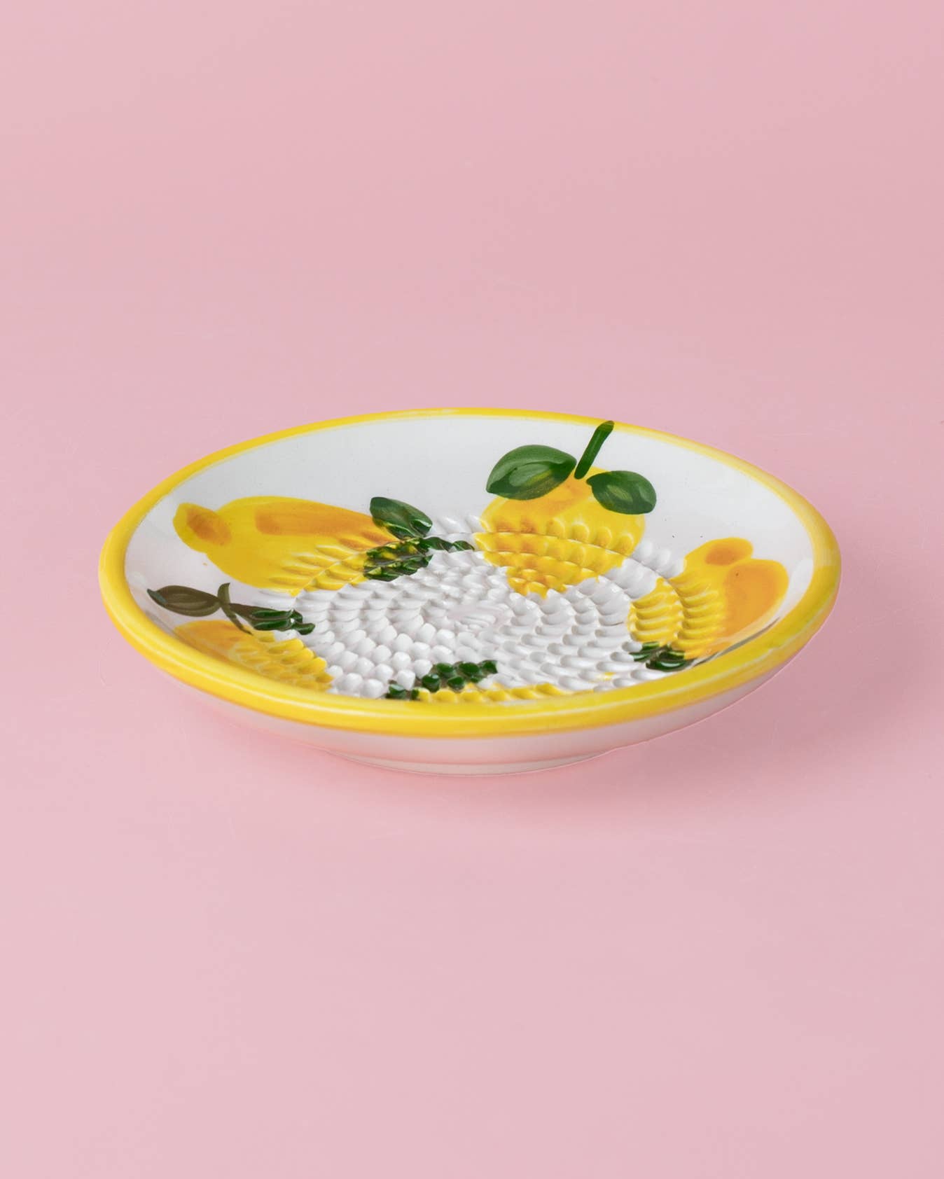 Die Alfar Tierra Cocida | 'Limone' Keramik Knoblauchreibe (12,5 x 12,5 x 2,5 cm) ist rund, handbemalt mit gelben Zitronen, grünen Blättern und gelbem Rand. Die strukturierte Mitte liegt auf zartem rosa Hintergrund.