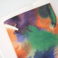 Eine Nahaufnahme des Papaya Paper Products | Notizheft A6 mit Aquarell/Watercolor-Motiv, mit einem leuchtenden Aquarell-Deckblatt in Grün, Orange, Lila und Blau und FSC-zertifizierten blanko Seiten, auf einer weißen Oberfläche.