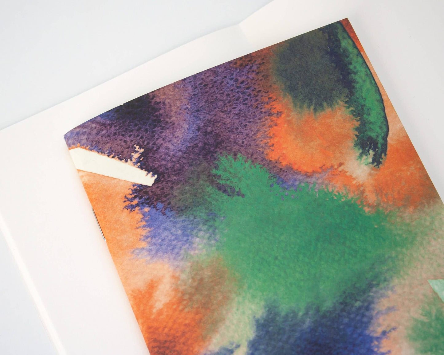 Eine Nahaufnahme des Papaya Paper Products | Notizheft A6 mit Aquarell/Watercolor-Motiv, mit einem leuchtenden Aquarell-Deckblatt in Grün, Orange, Lila und Blau und FSC-zertifizierten blanko Seiten, auf einer weißen Oberfläche.