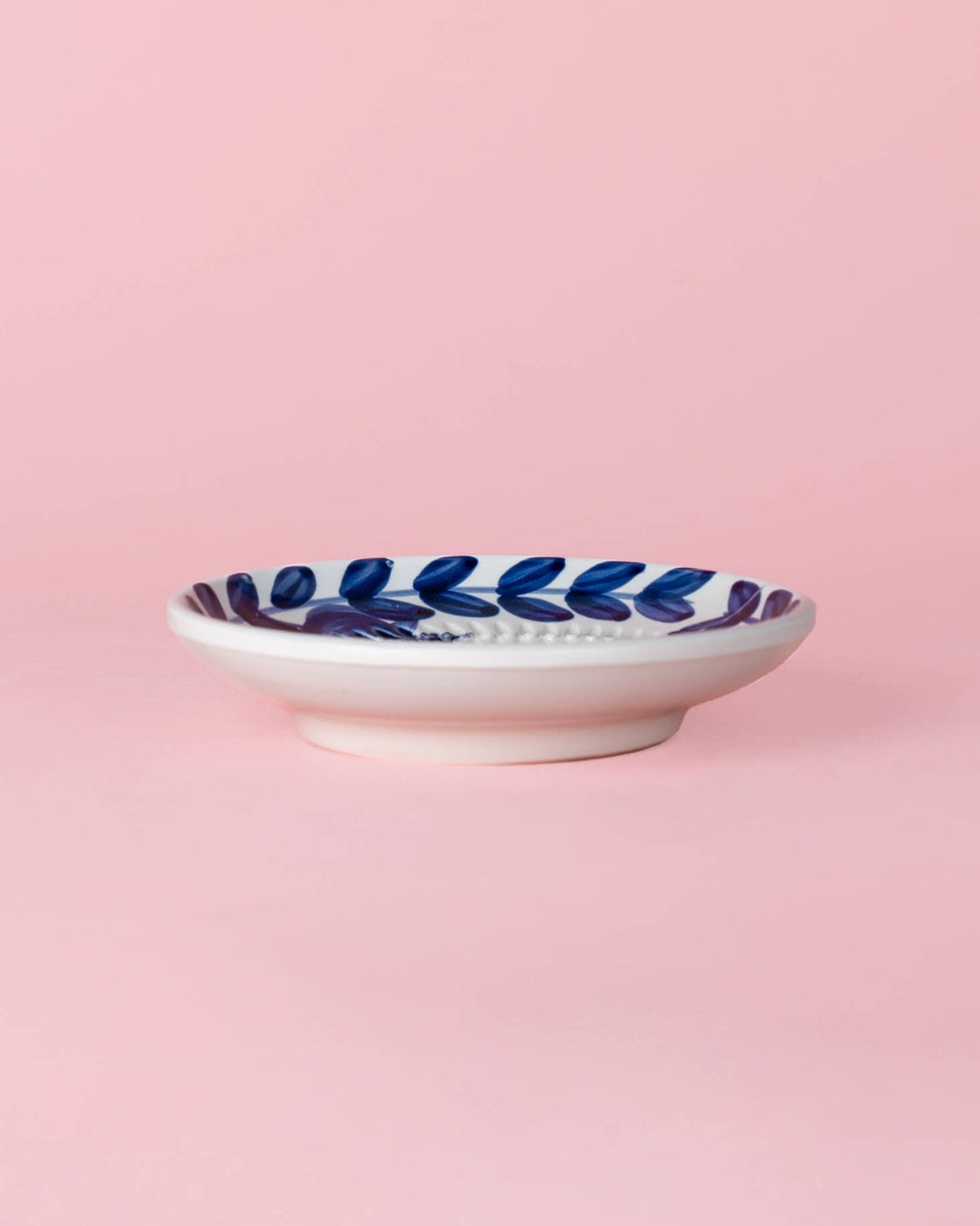 Eine kleine Keramik-Knoblauchreibplatte „Nerja“ von Alfar Tierra Cocida, traditionell von Hand in Blau bemalt und am Rand mit einem Blattmotiv verziert, präsentiert vor einem schlichten rosa Hintergrund.