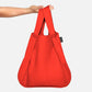 Eine Hand hält eine Notabag | „Original Red“ von Notabag – Tasche und Rucksack in einem – mit zwei Griffen vor weißem Hintergrund.