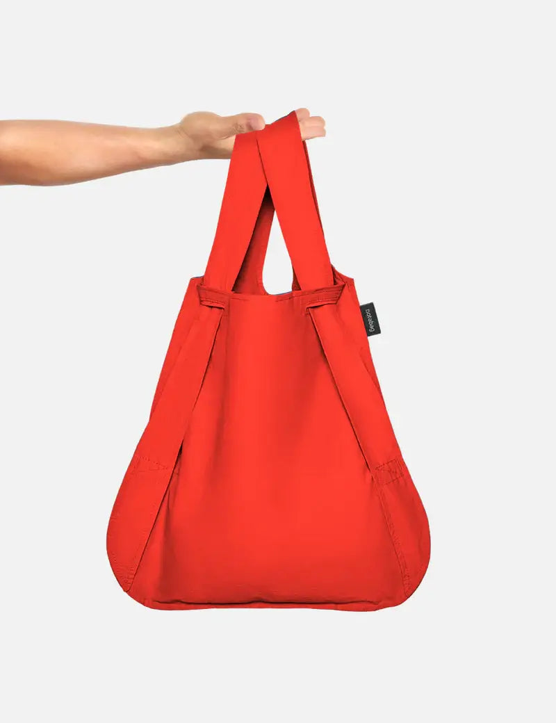Eine Hand hält eine Notabag | „Original Red“ von Notabag – Tasche und Rucksack in einem – mit zwei Griffen vor weißem Hintergrund.