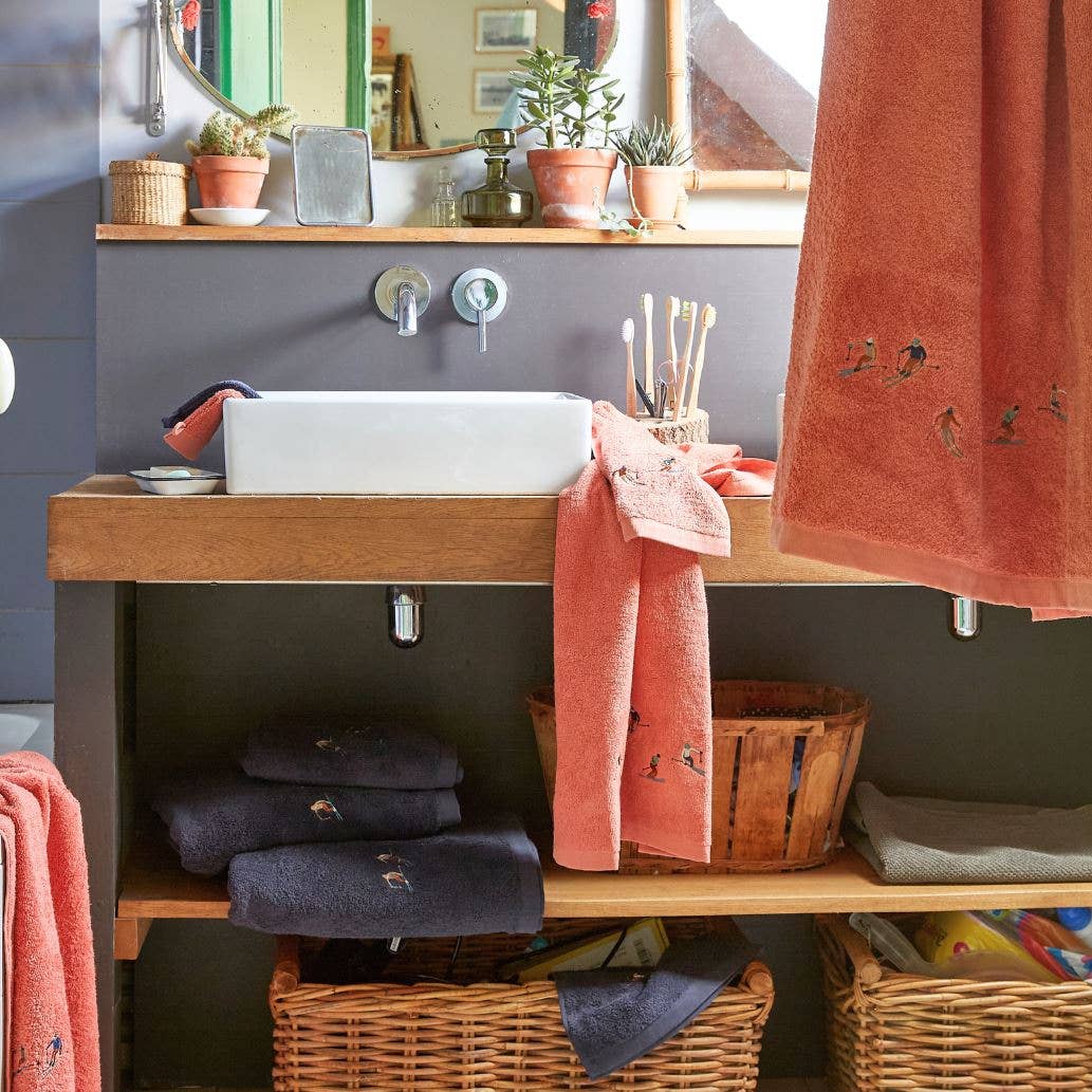 Das gemütliche Badezimmer ist mit einem modernen Waschbecken, korallen- und marineblauen Handtüchern, einem Spiegel, Topfpflanzen, OEKO-TEX-zertifizierten Sylvie Thiriez Waschlappen aus Frottee 'Après-Ski' Rosé, Zahnbürsten in einem Becher und geflochtenen Körben unter dem Waschbecken ausgestattet - alles fein säuberlich angeordnet.