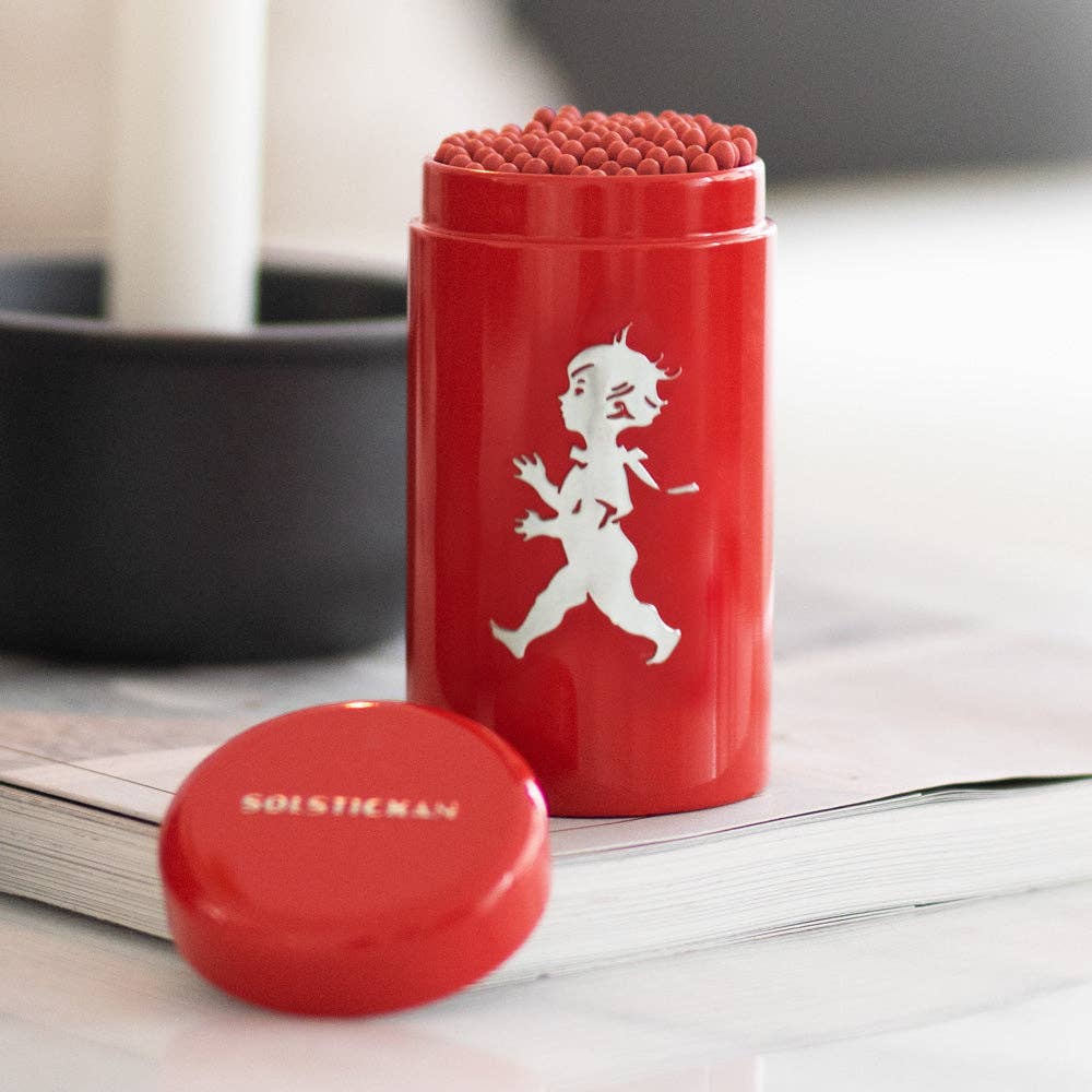 Der Streichholzhalter Solstickan Design | Streichholzdose Rot von Solstickan Design mit weißer Solsticke Boy-Silhouette, gefüllt mit extra langen Streichhölzern, steht auf einem Buch, daneben der Deckel SORI YANAGI.