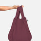 Eine Hand hält die Notabag | Wandelbare Tasche und Rucksack „Pflaume“ von Notabag – eine wiederverwendbare Tasche in einem satten Pflaumenton mit einem dezenten geometrischen Gittermuster und einem schwarz-weißen Seitenetikett –, die vor einem schlichten weißen Hintergrund präsentiert wird.