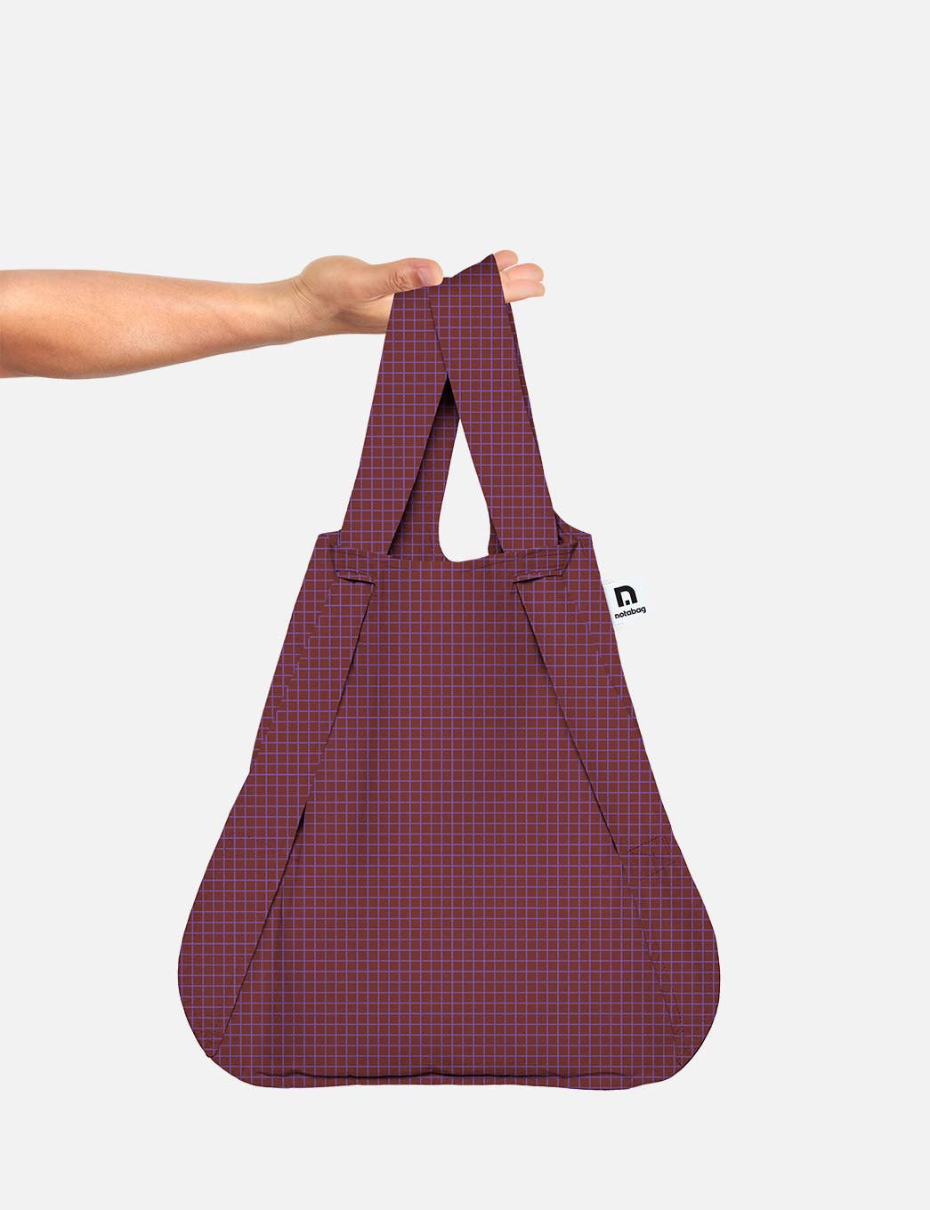 Eine Hand hält die Notabag | Wandelbare Tasche und Rucksack „Pflaume“ von Notabag – eine wiederverwendbare Tasche in einem satten Pflaumenton mit einem dezenten geometrischen Gittermuster und einem schwarz-weißen Seitenetikett –, die vor einem schlichten weißen Hintergrund präsentiert wird.