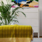 Ein modernes Wohnzimmer mit der gelben Lammfelldecke "Darling" von Sofie Sjöström über dem Sofa, einer Topfpflanze, einer weißen Kommode und einem farbenfrohen abstrakten Gemälde von Sofie Sjöström Design an der Wand.