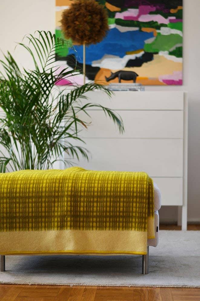 Ein modernes Wohnzimmer mit der gelben Lammfelldecke "Darling" von Sofie Sjöström über dem Sofa, einer Topfpflanze, einer weißen Kommode und einem farbenfrohen abstrakten Gemälde von Sofie Sjöström Design an der Wand.