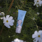 Eine Tube Archivist | 'Kokosnuss' Handcreme von Archivist Gallery mit einem illustrierten Gesicht auf der Verpackung steht aufrecht zwischen grünem Laub und weißen Blumen und bietet eine vegane Formel mit einem erfrischenden Kokosnussduft.