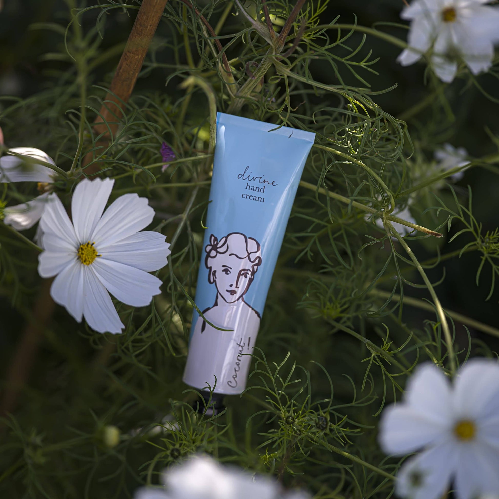 Eine Tube Archivist | 'Kokosnuss' Handcreme von Archivist Gallery mit einem illustrierten Gesicht auf der Verpackung steht aufrecht zwischen grünem Laub und weißen Blumen und bietet eine vegane Formel mit einem erfrischenden Kokosnussduft.