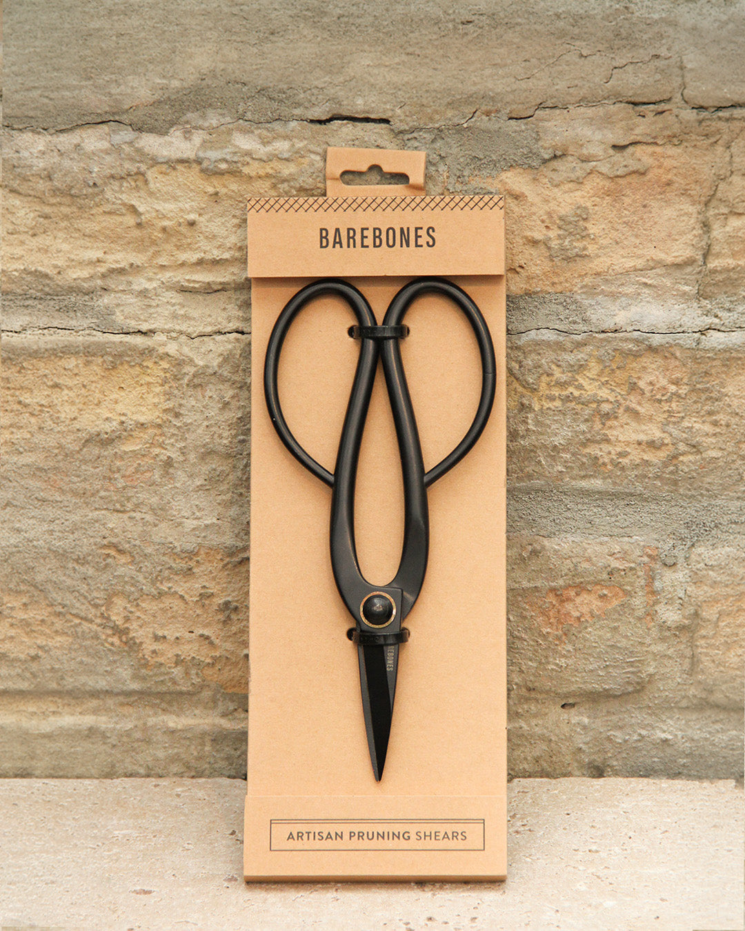 Schwarze Barebones | Artisan Gartenschere aus Edelstahl in einer minimalistischen Verpackung mit der Aufschrift Barebones; unten steht Artisan Pruning Shears" vor einem Hintergrund aus strukturiertem Stein.
