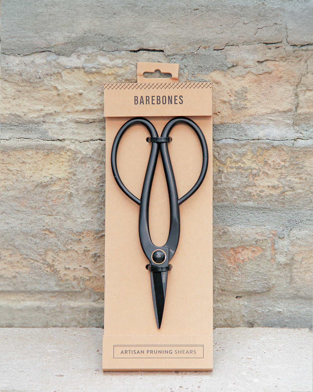 Schwarze Metall-Gartenscheren mit großen Griffen werden in einer minimalistischen braunen Verpackung mit der Aufschrift „Barebones“ und „Artisan Gartenschere aus Edelstahl“ vor einem strukturierten Steinhintergrund präsentiert – diese Barebones-Scheren verbinden elegantes Design mit professioneller Funktionalität.