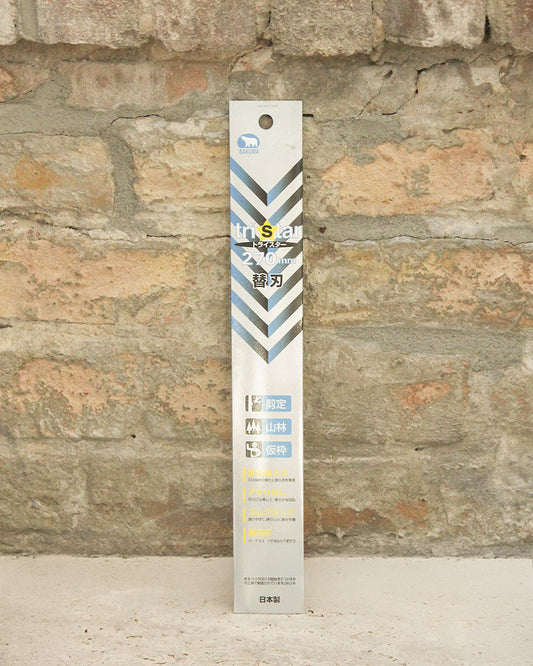 Eine rechteckige Schachtel mit blauem, grauem und weißem Chevron-Muster mit Niwaki-Schriftzug und zweisprachigem Text steht aufrecht vor einer rustikalen Ziegelwand. Das Produkt ist das Niwaki | Ersatzsägeblatt für Niwaki GR 270.