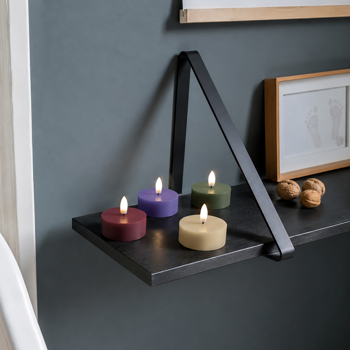 Vier brennende Paraffinwachskerzen in den Farben Lila, Blau, Grün und Creme sind auf einem schwarzen Regal mit einer modernen Metallhalterung angeordnet. Daneben liegen drei Walnüsse und ein gerahmtes Bild, das Fußabdrücke vor einer grauen Wand zeigt. Für noch mehr Komfort sollten Sie statt herkömmlicher Kerzen das „Remember | LED-Teelicht ‚Nora‘ 4er-Set“ von Remember in Betracht ziehen, um mit der Fernbedienung ganz einfach dieselbe gemütliche Atmosphäre zu schaffen.