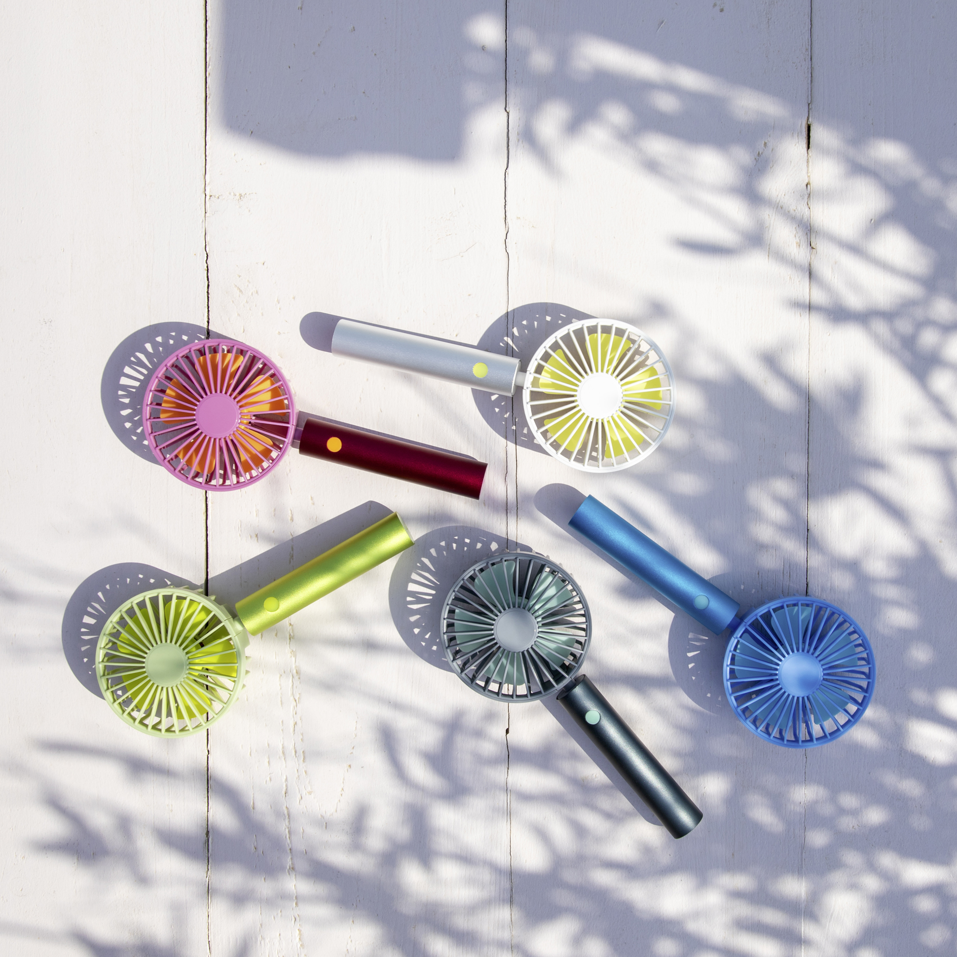 Sechs Remember 'Flow Aqua'-Ventilatoren sind auf einer weißen Holzfläche angeordnet, wobei Sonnenlicht und Blätterschatten eine lebendige, sommerliche Atmosphäre schaffen und die bunten Ventilatoren von Remember hervorheben.