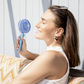 Eine Frau mit braunen Haaren, geschlossenen Augen und lächelnd, sitzt auf einer gemusterten Couch und hält ihren Remember Ventilator 'Flow Aqua' von Remember an ihr Gesicht. Das Sonnenlicht scheint auf sie und die Sonnenbrille ruht auf ihrem Kopf.