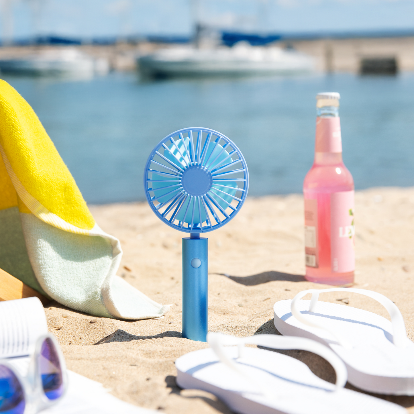Der Remember Ventilator 'Flow Aqua' steht aufrecht auf einem Sandstrand neben einem Handtuch, einer rosa Limonadenflasche, weißen Flip-Flops und einer Sonnenbrille, mit verschwommenen Booten und aquatischem Wasser im Hintergrund.