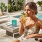 Eine Frau in einem leichten Sommerkleid entspannt sich am Pool und genießt einen Eistee von English Tea Shop | Tropical Mint, BIO, 20 Teebeutel by Royal Nature GmbH & Co. KG. Ihr Smartphone und eine Zeitschrift liegen auf dem Tisch neben ihr.