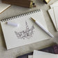 Ein Skizzenbuch mit Gebäuden und einem Kirchturm liegt auf einem Schreibtisch, umgeben von Kunstzubehör wie dem ENTREPÔT YSTUDIO Harz-Rollerball-Stift (Weiß), einem Stift, Radiergummi und Papier.