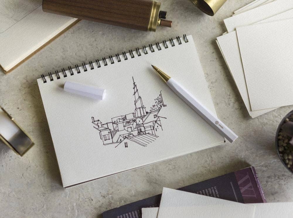 Ein Skizzenbuch mit Gebäuden und einem Kirchturm liegt auf einem Schreibtisch, umgeben von Kunstzubehör wie dem ENTREPÔT YSTUDIO Harz-Rollerball-Stift (Weiß), einem Stift, Radiergummi und Papier.