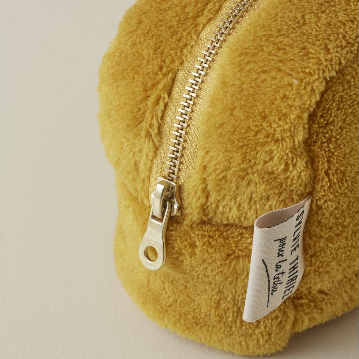 Nahaufnahme der Sylvie Thiriez Kosmetiktasche Honigfarben aus Plüsch mit goldenem Reißverschluss und weißem Seitenetikett. Diese weiche Tasche von Sylvie Thiriez ist OEKO-TEX zertifiziert und liegt auf einer leichten Oberfläche.