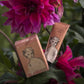 Eine Tube und eine Schachtel der "Kirschblüte" Handcreme von Archivist Gallery mit illustrierten Frauengesichtern liegen inmitten von leuchtend rosafarbenen Blumen und üppig grünem Laub.