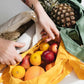 Eine Person mit einem Tattoo auf dem Arm packt Lebensmittel – Ananas, Äpfel, Orangen und ein Baguette – in die Notabag | „Original Golden“, eine vielseitige Tragetasche und Rucksack von Notabag.