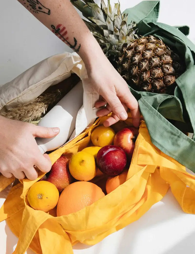 Eine Person mit einem Tattoo auf dem Arm packt Lebensmittel – Ananas, Äpfel, Orangen und ein Baguette – in die Notabag | „Original Golden“, eine vielseitige Tragetasche und Rucksack von Notabag.