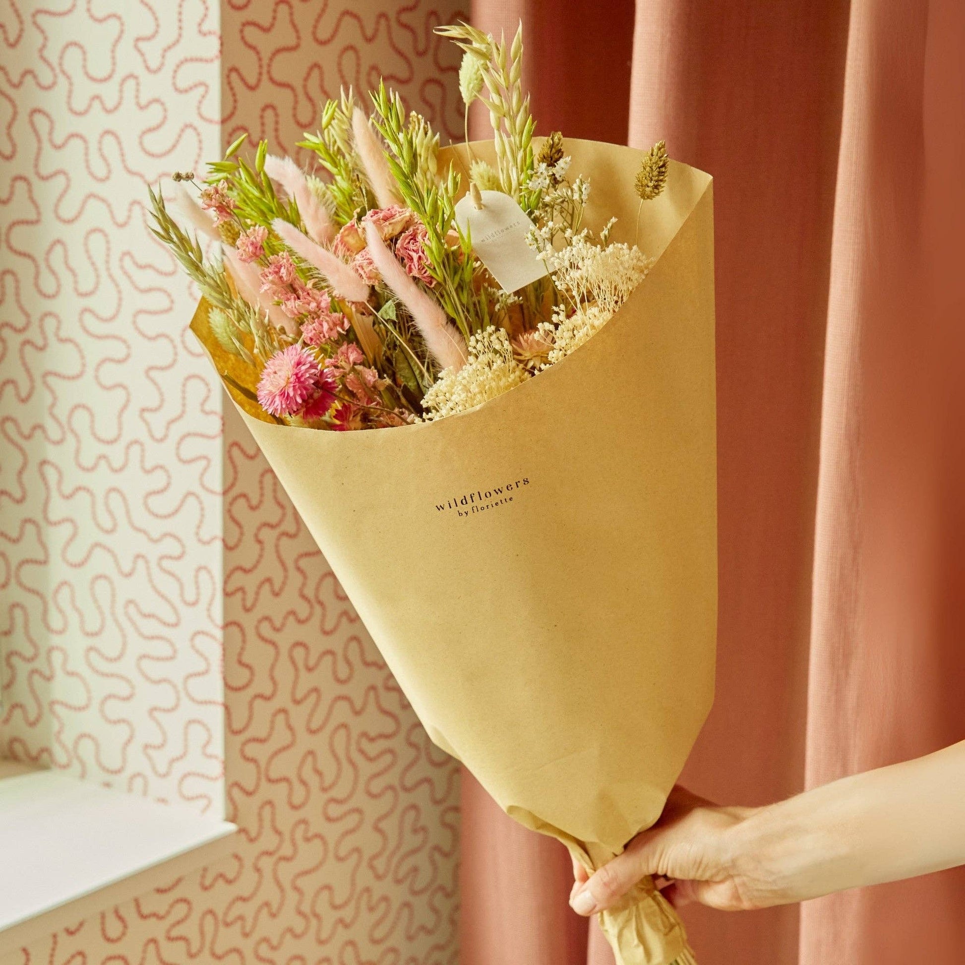 Eine Hand hält den Feldstrauß „Rosa Eleganz“ in Größe S von Wildflowers by Floriette – einen bezaubernden Strauß aus getrockneten Wildblumen, eingewickelt in braunes Papier, vor dem Hintergrund eines rosa Vorhangs und einer gemusterten Tapete – eine Szene, die pure „Rosa Eleganz“ ausstrahlt.