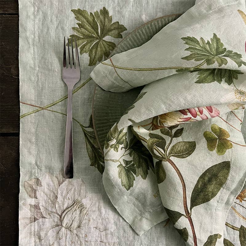 Eine Leinenserviette „Flora Danica Atlas“ von Koustrupco (45 × 45 cm) liegt zusammen mit einer Gabel neben einem passenden Teller mit botanischem Motiv – beide mit grünen Blatt- und Blumenmustern – auf einem dunklen Holztisch.