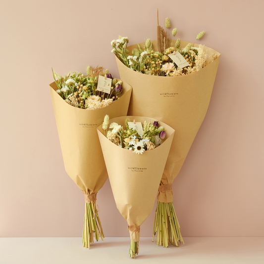 Drei Blumensträuße „Wildflowers by Floriette | Getrockneter Feldblumenstrauß ‚Natur‘ in M“, arrangiert vor einem hellrosa Hintergrund, jeweils in braunes Papier eingewickelt, an den Stielen zusammengebunden und mit einer kleinen Karte verziert.