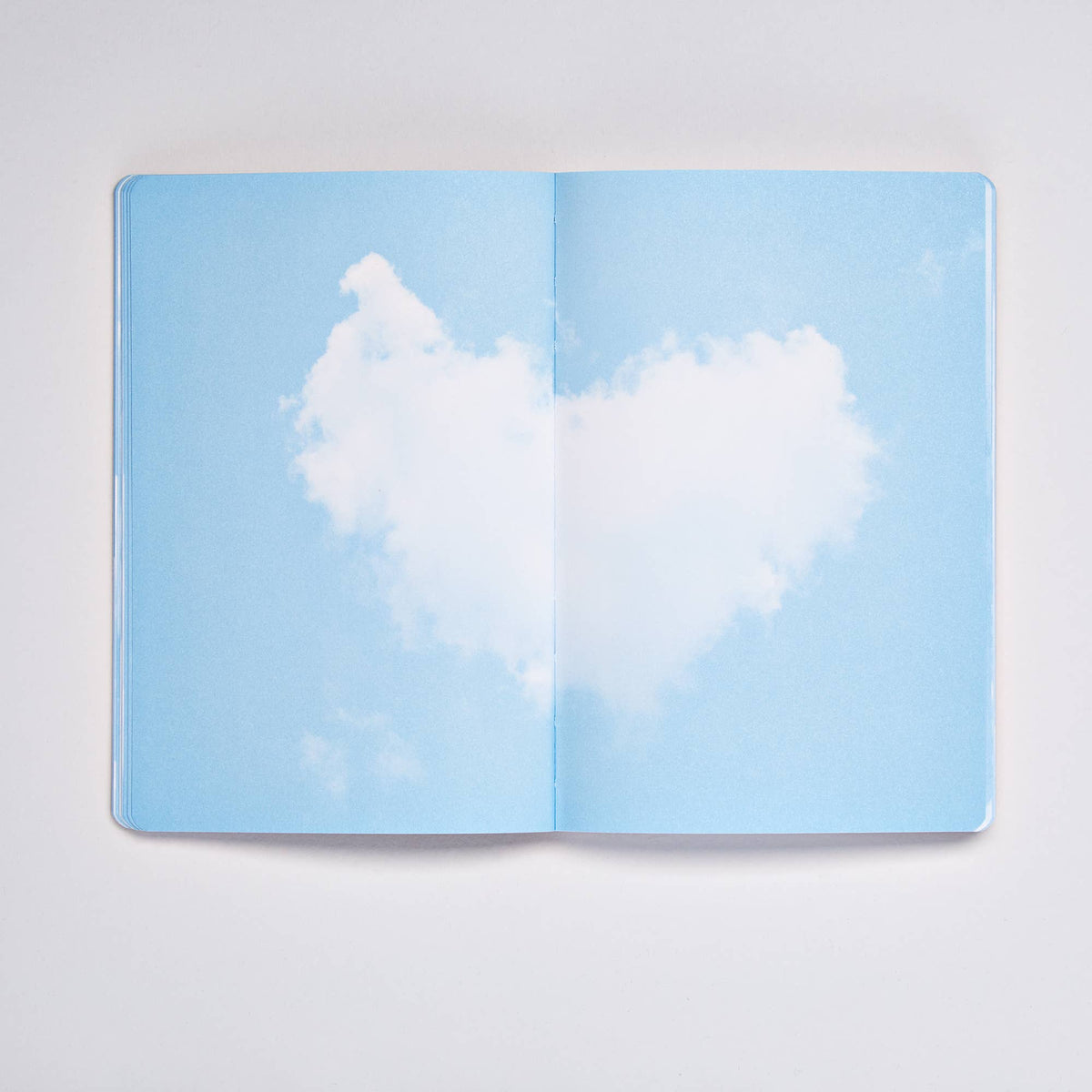 Ein aufgeschlagenes Buch zeigt eine herzförmige weiße Wolke, die vor einem blauen Himmel schwebt; das Bild erstreckt sich nahtlos über beide Seiten – ein verträumtes „Cloud Blue“-Motiv von nuuna, das Inspiration und Fantasie beflügelt. Dieses wunderschön gestaltete nuuna | „Cloud Blue“-Inspirationsbuch in der Größe M mit 176 farbenfrohen Seiten und einem flexiblen Flexcover verwandelt jede Notiz und jede Skizze in etwas Außergewöhnliches.