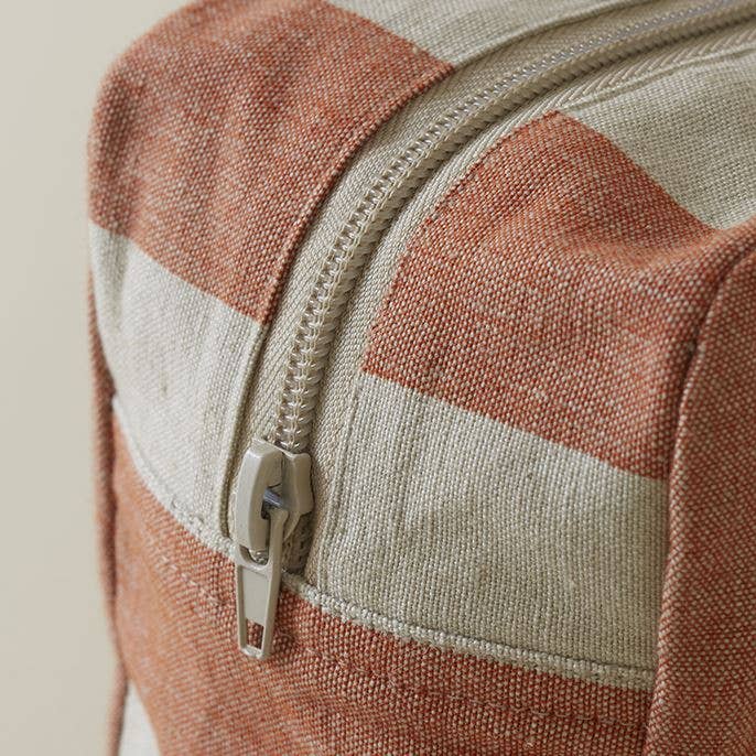 Nahaufnahme der Sylvie Thiriez Kosmetiktasche 'Mistral' klein: eine kompakte Kosmetiktasche aus Stoff in ziegelrot, mit breiten horizontalen Streifen in Orange und Off-White, strukturiertem Material und einem Metallreißverschluss.