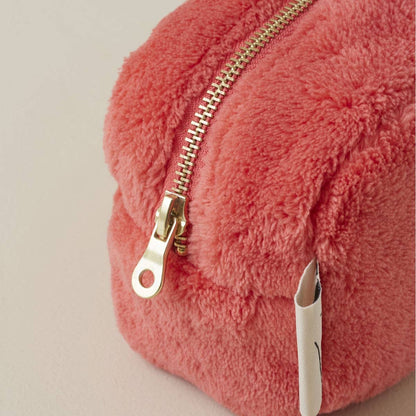 Eine Nahaufnahme der Sylvie Thiriez | Kosmetiktasche Flamingofarben aus Plüsch, einer zartrosa Tasche mit goldenem Reißverschluss und weißem Stoffanhänger von Sylvie Thiriez, die auf einer hellen Oberfläche ruht.