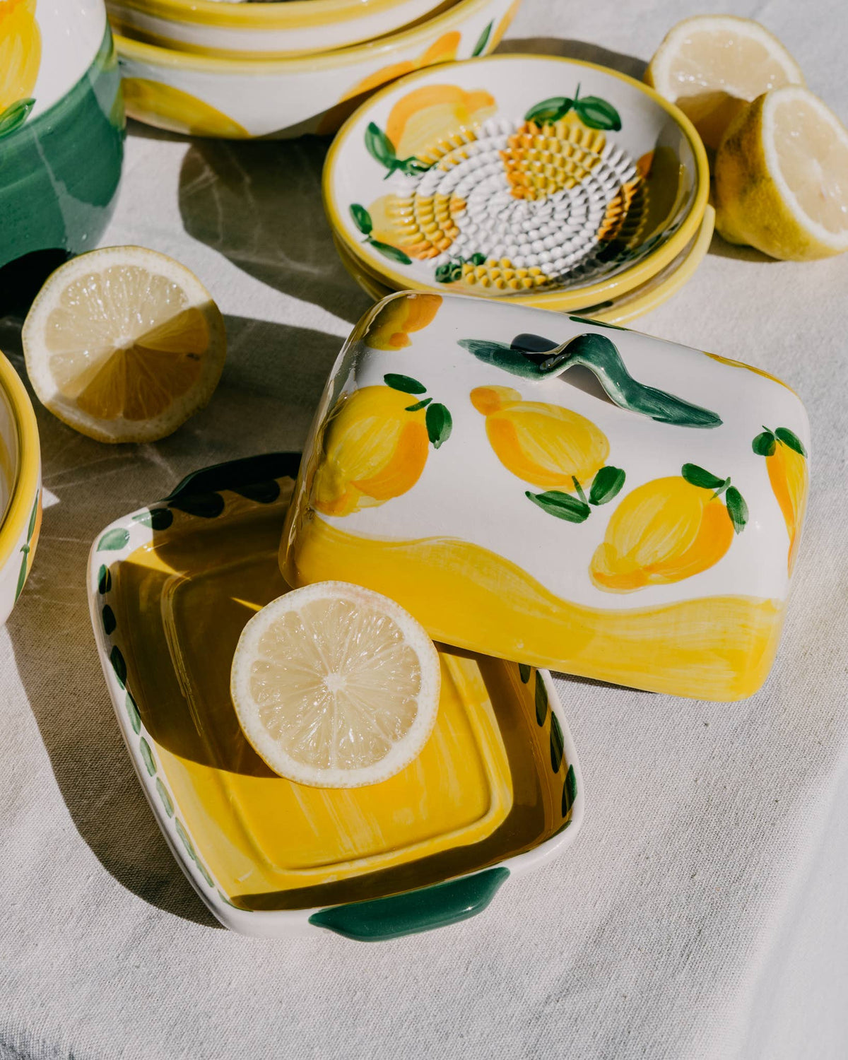 Handgefertigtes Keramikgeschirr von Alfar Tierra Cocida schmückt den Tisch: Im Mittelpunkt steht die handgefertigte Keramik-Butterdose „Limone“ von Alfar Tierra Cocida in Gelb und Weiß, geöffnet und mit einer Zitronenscheibe gefüllt. Umgeben von mediterranen Tellern und halbierten Zitronen auf einer hellen Tischdecke kommen die handbemalten Details der Keramik im natürlichen Licht besonders schön zur Geltung und schaffen eine frische Atmosphäre.