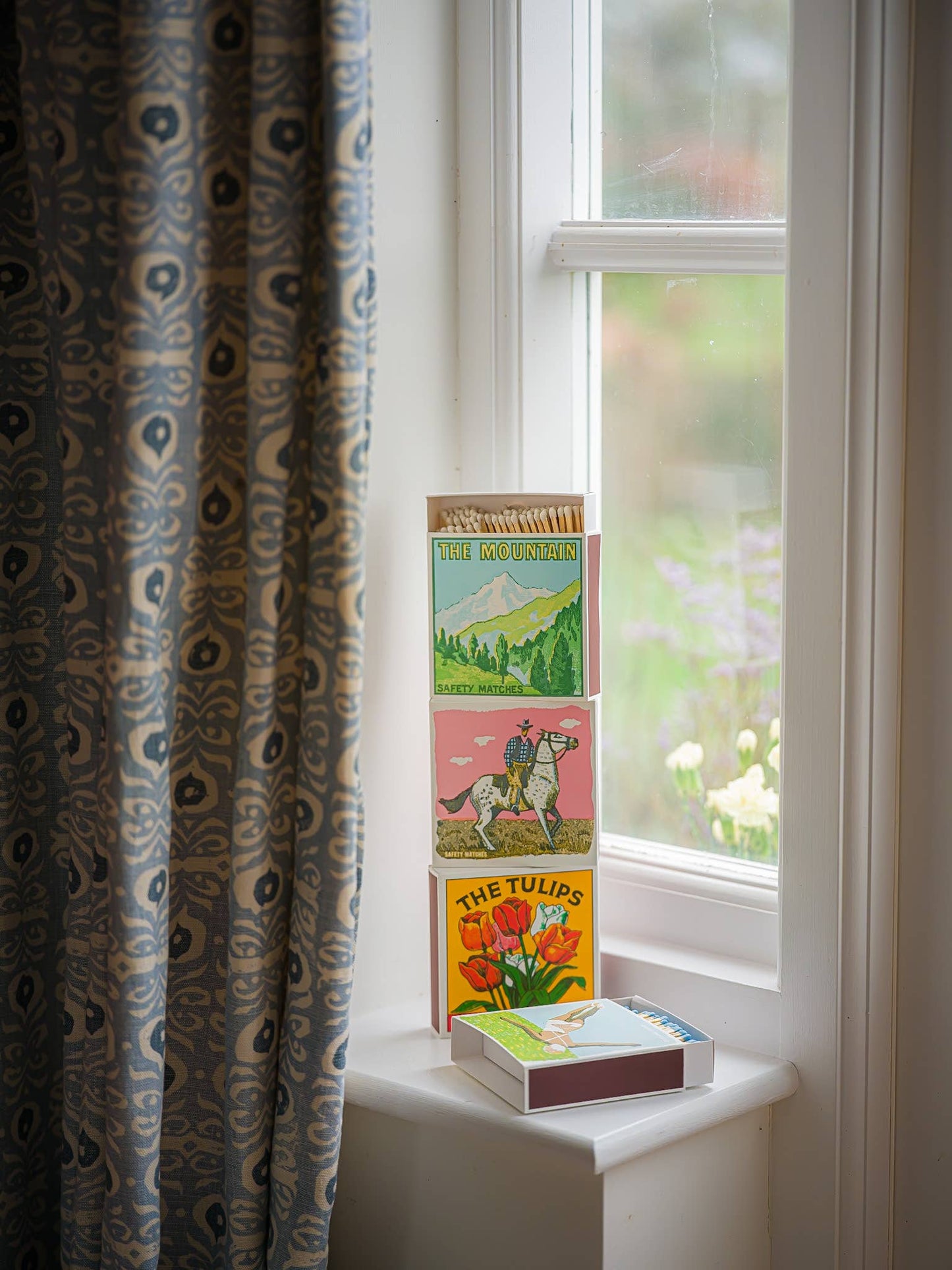 Drei bunte Bücher stapeln sich auf einer weißen Fensterbank neben einem gemusterten Vorhang. In der Nähe ruht Archivist Gallery's "Die Tulpen" Quadratische Streichhölzer, während Grün und Blumen außerhalb des Fensters im Weichzeichner erscheinen.