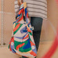 Eine Person hält eine Notabag | 'Flow' Kombination aus Tasche & Rucksack von Notabag, die abstrakte rote, grüne, blaue und orangefarbene Wirbel aufweist. Sie trägt eine gestreifte Tunika, dunkle Hosen und schwarze Schuhe; im Hintergrund ist eine perforierte Wand zu sehen.