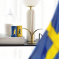 Nahaufnahme eines Tisches mit einer schwedischen Flagge, zwei Streichholzschachteln von Solstickan Design Streichholzdose Gelb, einer weißen Kerze in einem goldenen Halter und einem Stapel Bücher, auf dem eine weiße Tasse unter einer Glaskuppel steht.