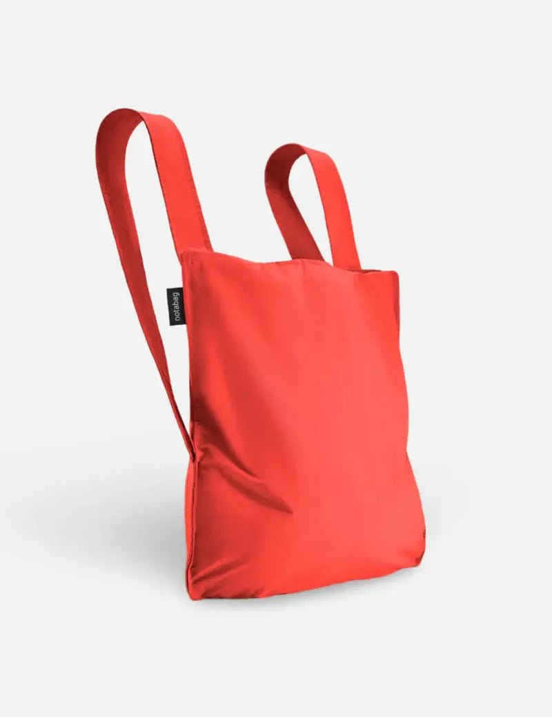 Der Notabag | „Original Red“ – Tasche und Rucksack in einem von Notabag – steht mit zwei Tragegriffen aufrecht und vereint die Funktionalität eines Rucksacks und einer Tragetasche in einer umweltfreundlichen, stilvollen Mehrwegtasche vor einem schlichten weißen Hintergrund.