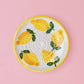Die Alfar Tierra Cocida 'Limone' Keramik Knoblauchreibe (12,5 x 12,5 x 2,5 cm) ist ein runder, handbemalter Keramikteller mit Zitronenmotiv und grünen Blättern, der in der Mitte eine erhabene Struktur zum Reiben aufweist und auf einem rosa Hintergrund abgebildet ist.