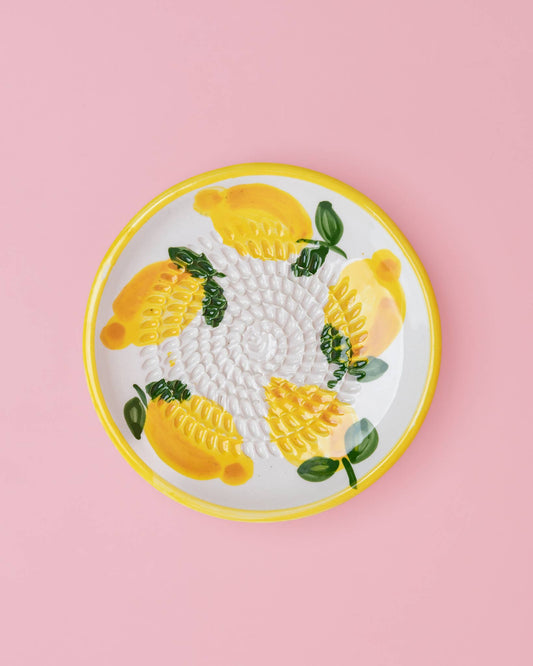 Die Alfar Tierra Cocida 'Limone' Keramik Knoblauchreibe (12,5 x 12,5 x 2,5 cm) ist ein runder, handbemalter Keramikteller mit Zitronenmotiv und grünen Blättern, der in der Mitte eine erhabene Struktur zum Reiben aufweist und auf einem rosa Hintergrund abgebildet ist.