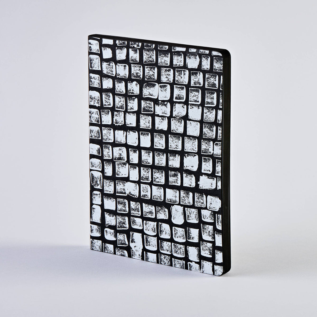 Ein nuuna | „Sassy Squares“ Notizbuch Not Ordinary L Light A5+ steht aufrecht vor einem schlichten, hellen Hintergrund; sein schwarzer, strukturierter Einband ist mit einem markanten Raster aus unregelmäßigen weißen Quadraten verziert, das an Steinfliesen erinnert.