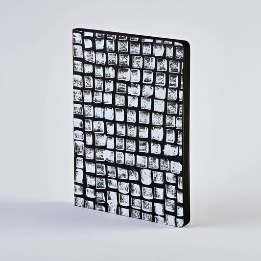 Ein nuuna | „Sassy Squares“ Notizbuch Not Ordinary L Light A5+ steht aufrecht vor einem schlichten, hellen Hintergrund; sein schwarzer, strukturierter Einband ist mit einem markanten Raster aus unregelmäßigen weißen Quadraten verziert, das an Steinfliesen erinnert.